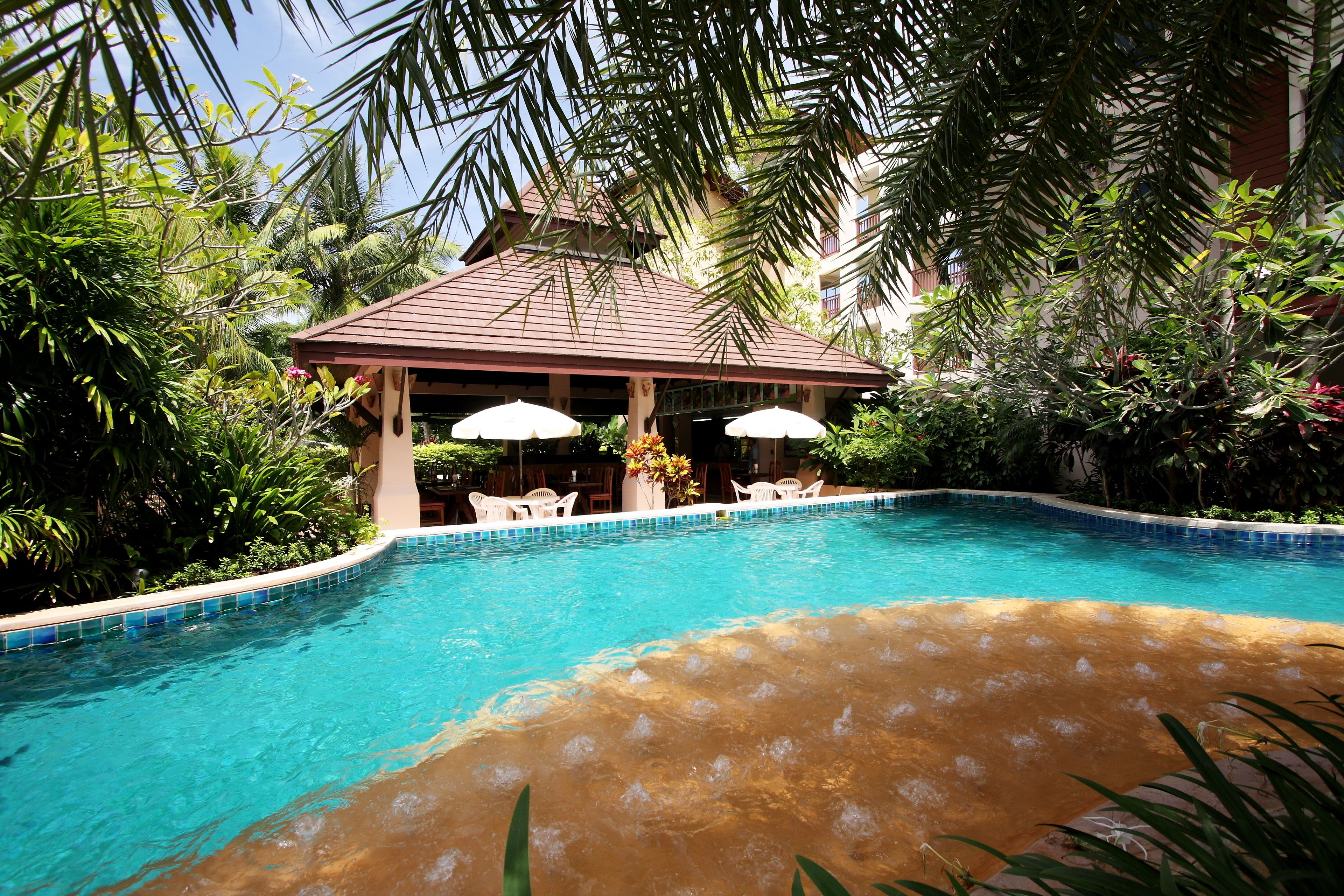 A swimming pool with a tiled edge, surrounded by lush greenery and a covered seating area with umbrellas.