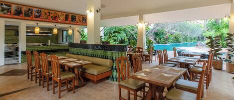 Un restaurante con una zona de bar de azulejos verdes, mesas y sillas de madera y vistas a una piscina.