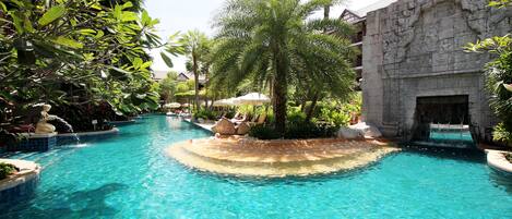 A pool area with clear water, surrounded by tropical plants and a stone building with a waterfall feature.