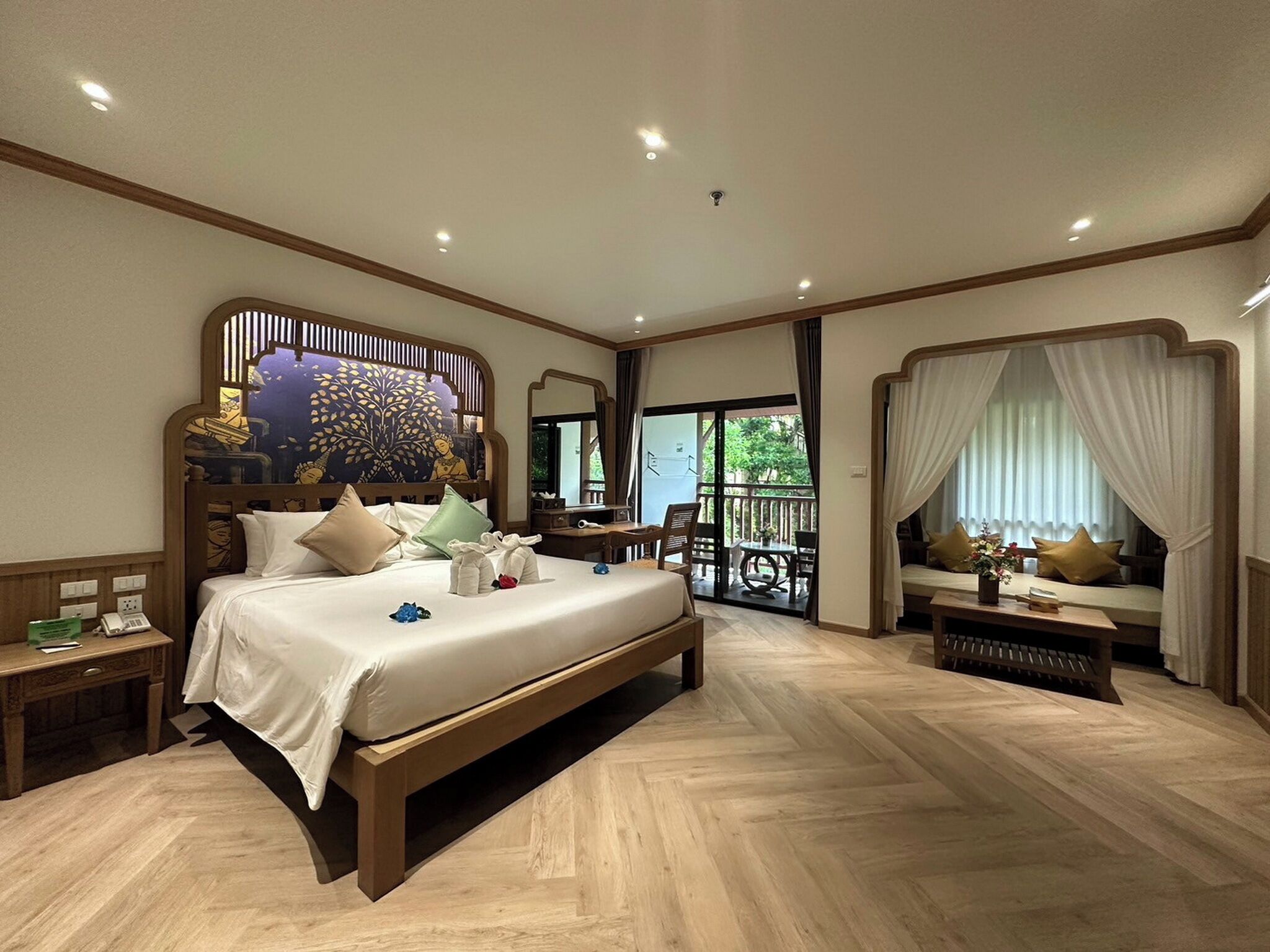 A spacious bedroom with a large bed, a wooden headboard, a desk, and a balcony with a view.