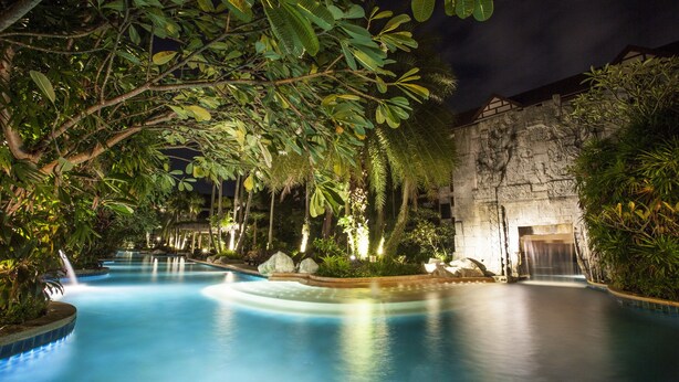 A nighttime swimming pool with a waterfall, surrounded by lush greenery and a stone wall.