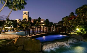 A bridge over a pool with a waterfall, surrounded by palm trees and a building with a tower. - Avanti Holiday Village (Paphos)