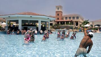 A swimming pool area with people engaging in water activities, a covered pavilion, and a building with a tower in the background.