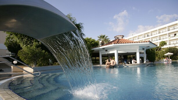 Une piscine agrémentée d’un élément décoratif aquatique, un pavillon couvert et un bâtiment doté de nombreux balcons en arrière-plan.
