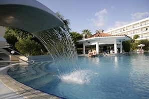 Ein Pool mit Wasserspiel, ein überdachter Pavillon und im Hintergrund ein Gebäude mit mehreren Balkonen.