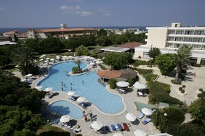 Ein großer Pool mit mehreren Liegebereichen und Sonnenschirmen, umgeben von üppiger Vegetation und Gebäuden.