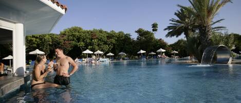 A swimming pool with tiled flooring, palm trees, and umbrellas in the background.