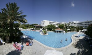 Ein großer Schwimmteich mit mehreren Bahnen, umgeben von Liegestühlen und Sonnenschirmen, und ein Hotelgebäude im Hintergrund.