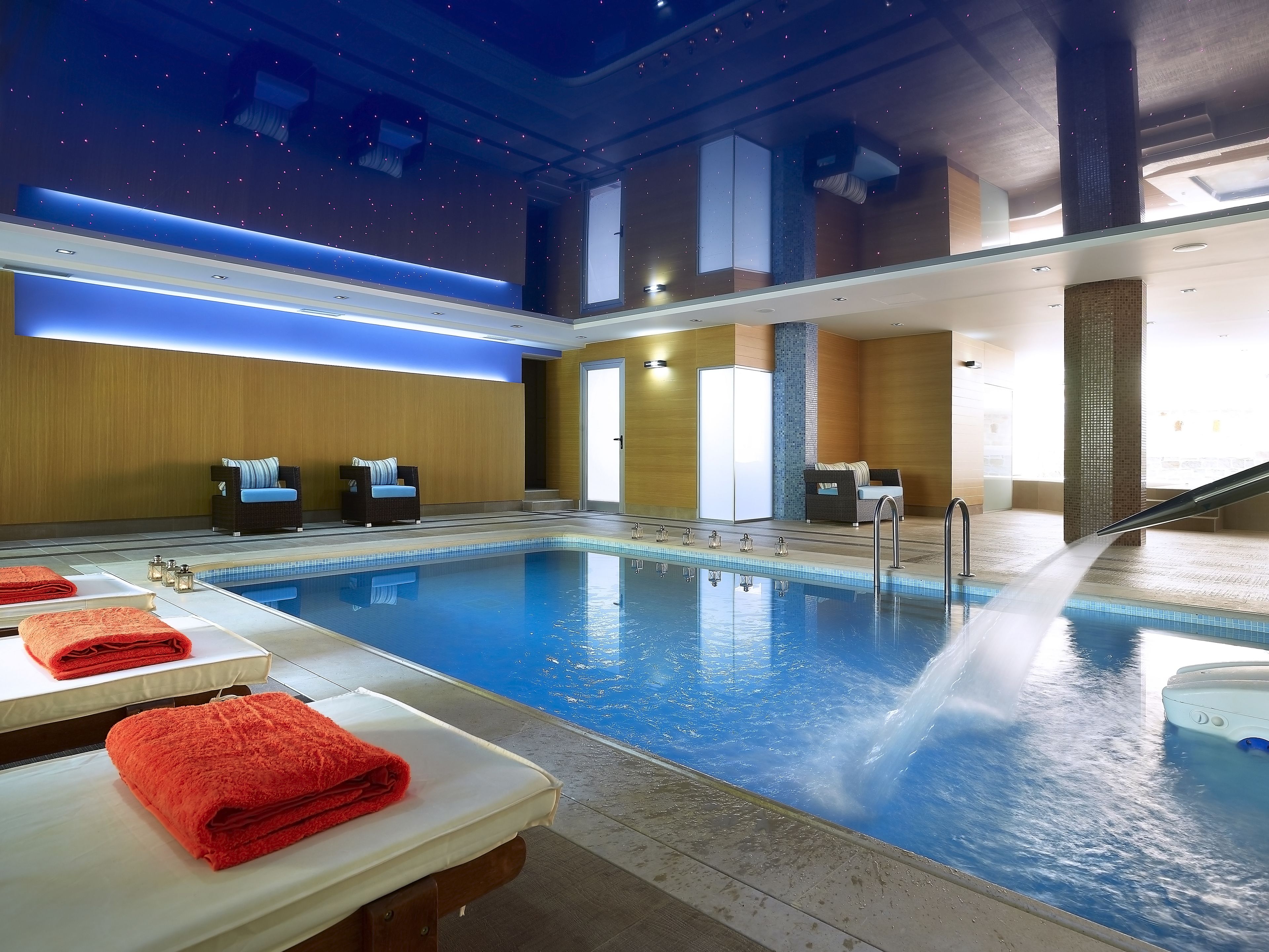 An indoor swimming pool with a water fountain, lounge chairs, and towels on tables.