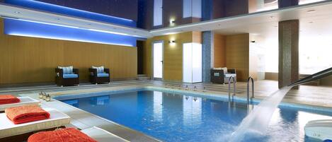 An indoor swimming pool with a water fountain, lounge chairs, and towels on tables.