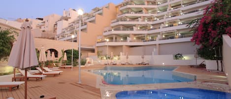 A pool area with lounge chairs and umbrellas, a multi-story building in the background, and a clear sky.