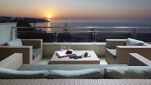 A balcony with wicker furniture, a table with a red vase, and a view of the sunset over the sea. - Macaris Suites and SPA (Rethymno)