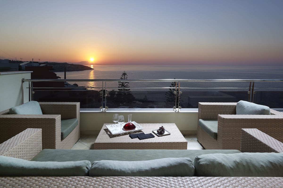 Un balcón con muebles de mimbre, una mesa con un florero rojo y vista al atardecer sobre el mar.