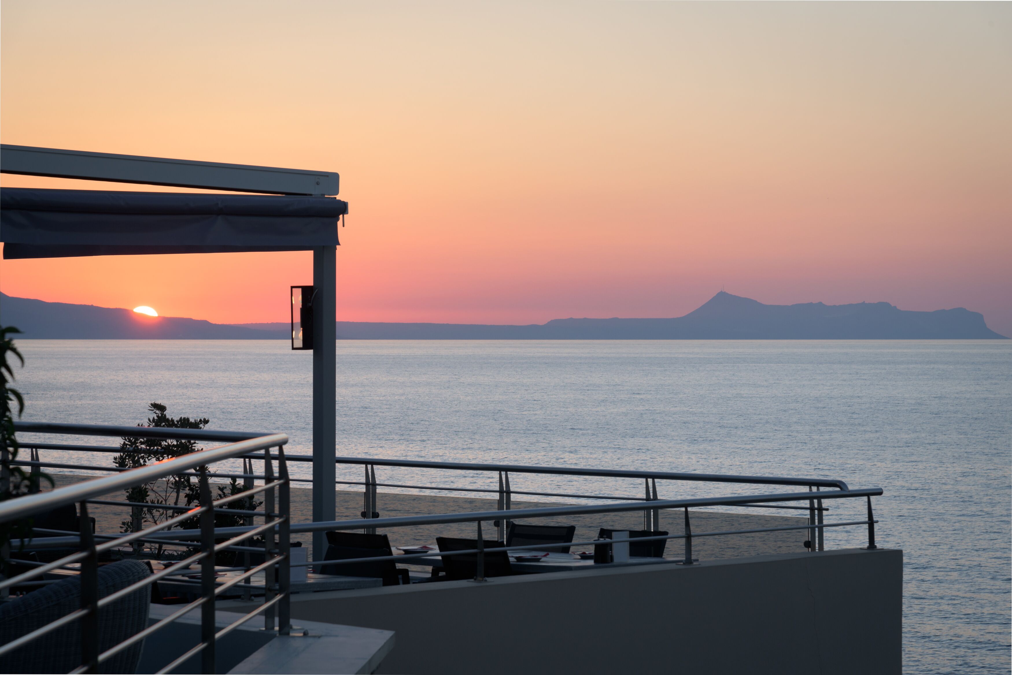 Una terraza con vistas a la puesta de sol sobre el mar, que incluye asientos al aire libre y una barandilla.