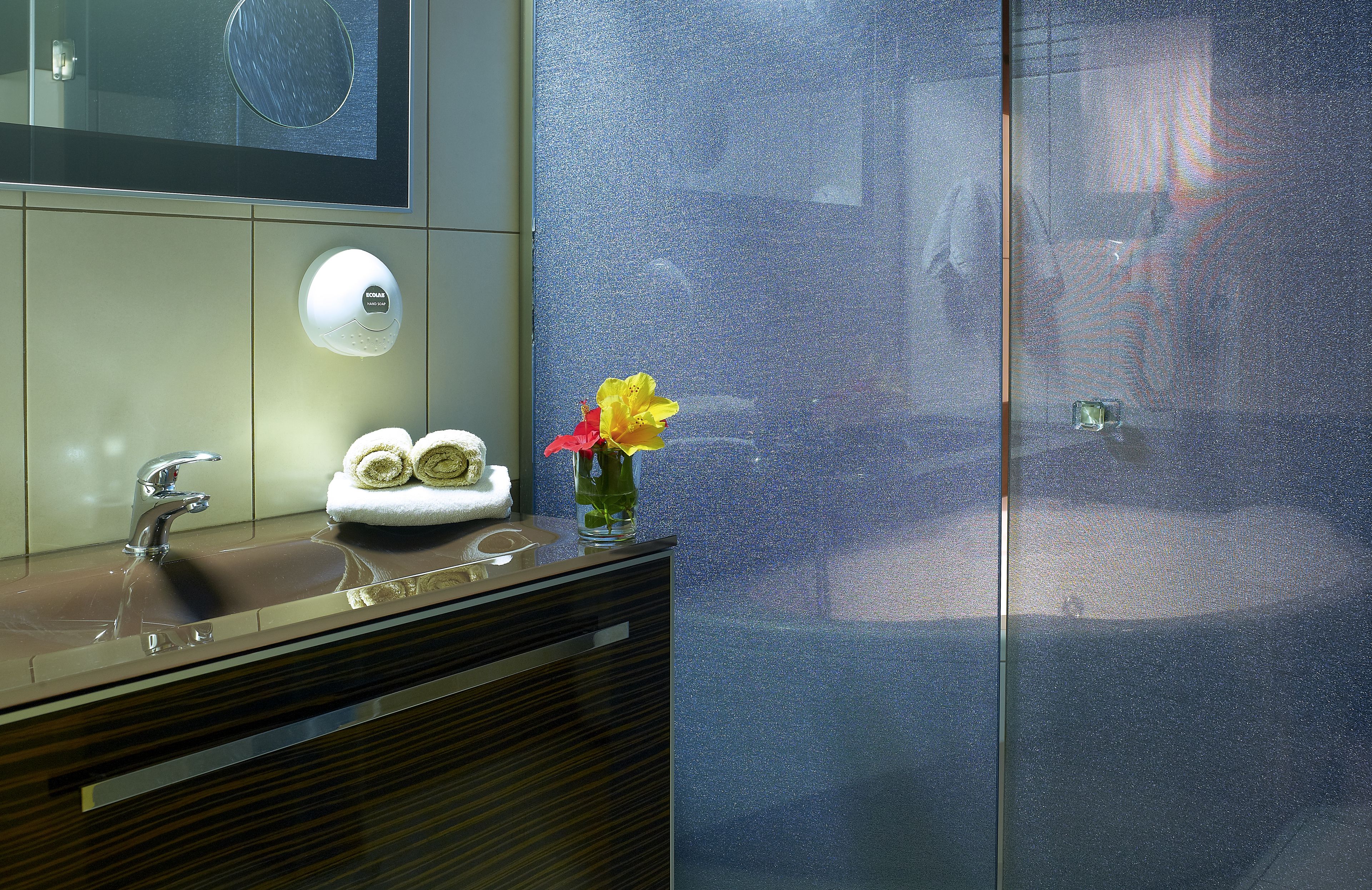 Une salle de bain moderne avec un grand miroir, un lavabo muni d’un porte-serviettes et un vase rempli de fleurs.