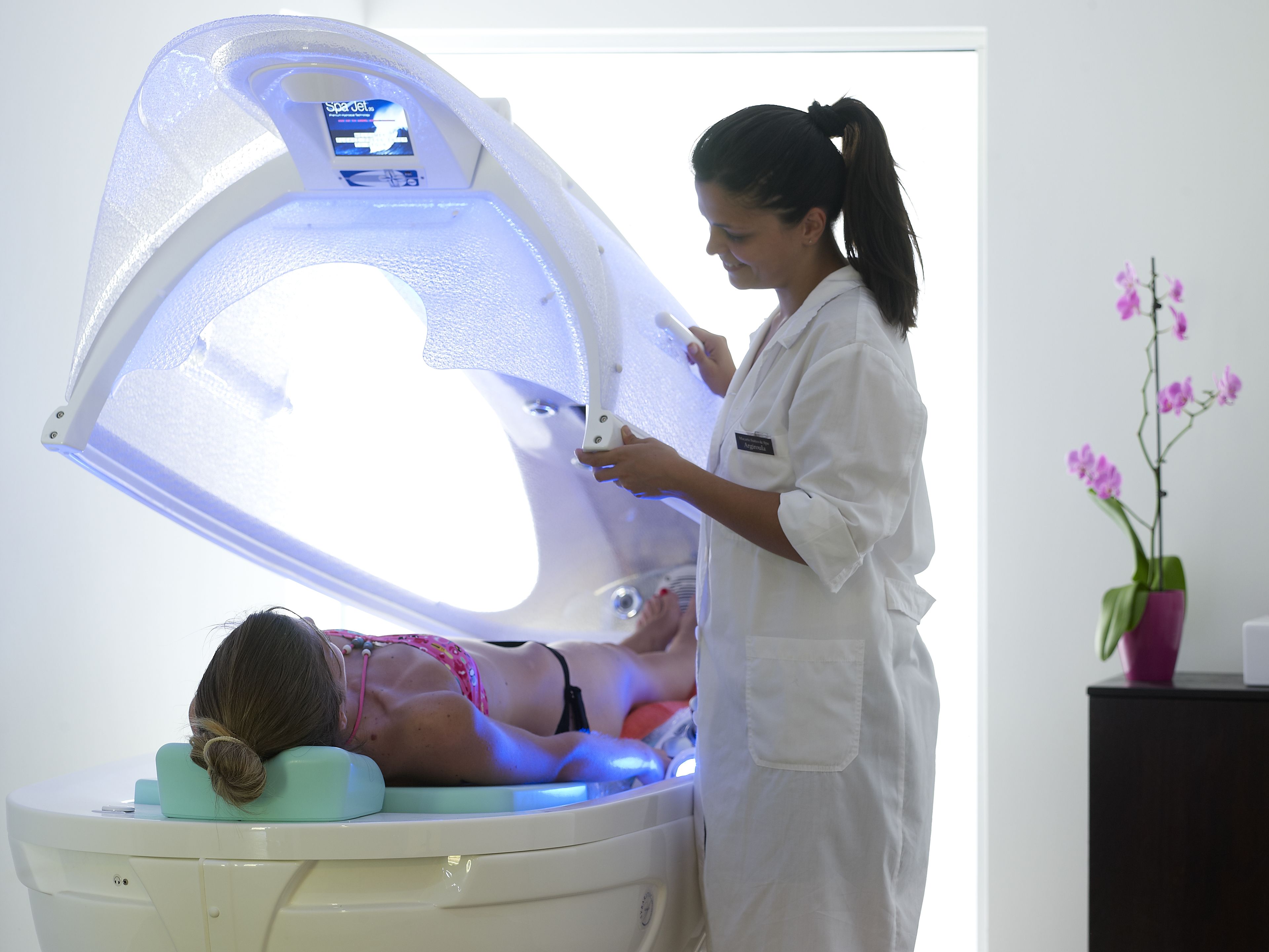A person lying in a treatment bed with a white cover, a woman standing beside it, and a pink orchid in a pot on a black surface.