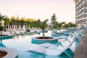 A pool area with multiple circular water features, lounge chairs, and palm trees. - Asterias Beach Hotel (Ayia Napa)