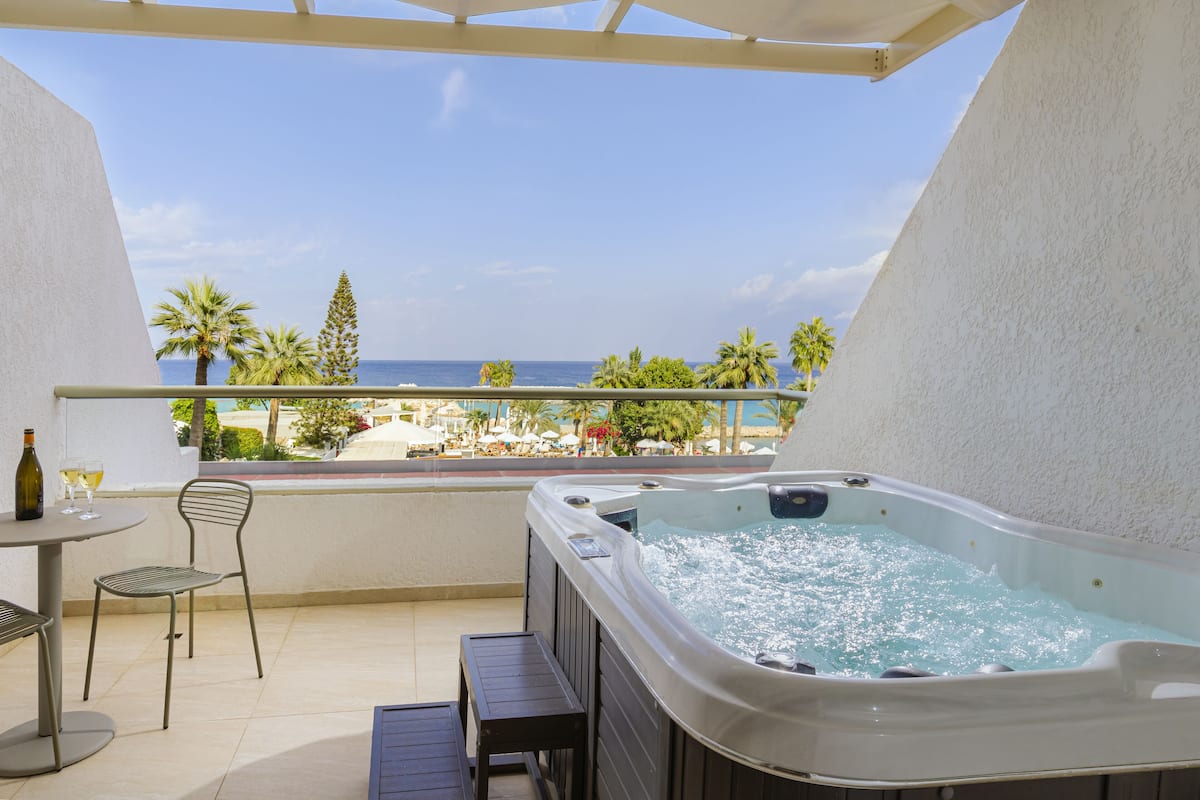 Un balcón con hidromasaje, una mesa con sillas y vista a la playa y palmeras.