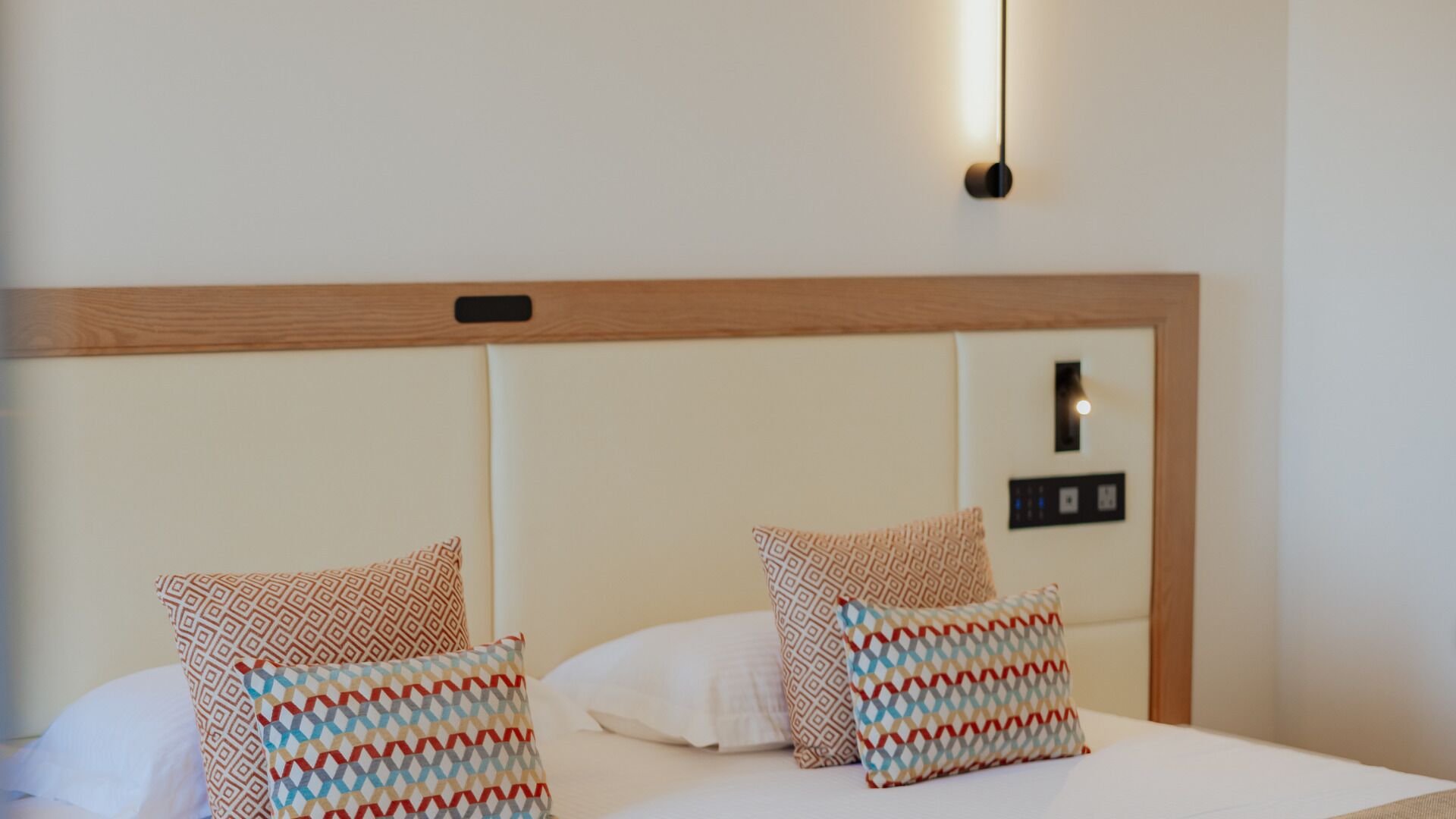 A hotel room with a wooden headboard, two patterned pillows, a white bedspread, and a wall-mounted light fixture.