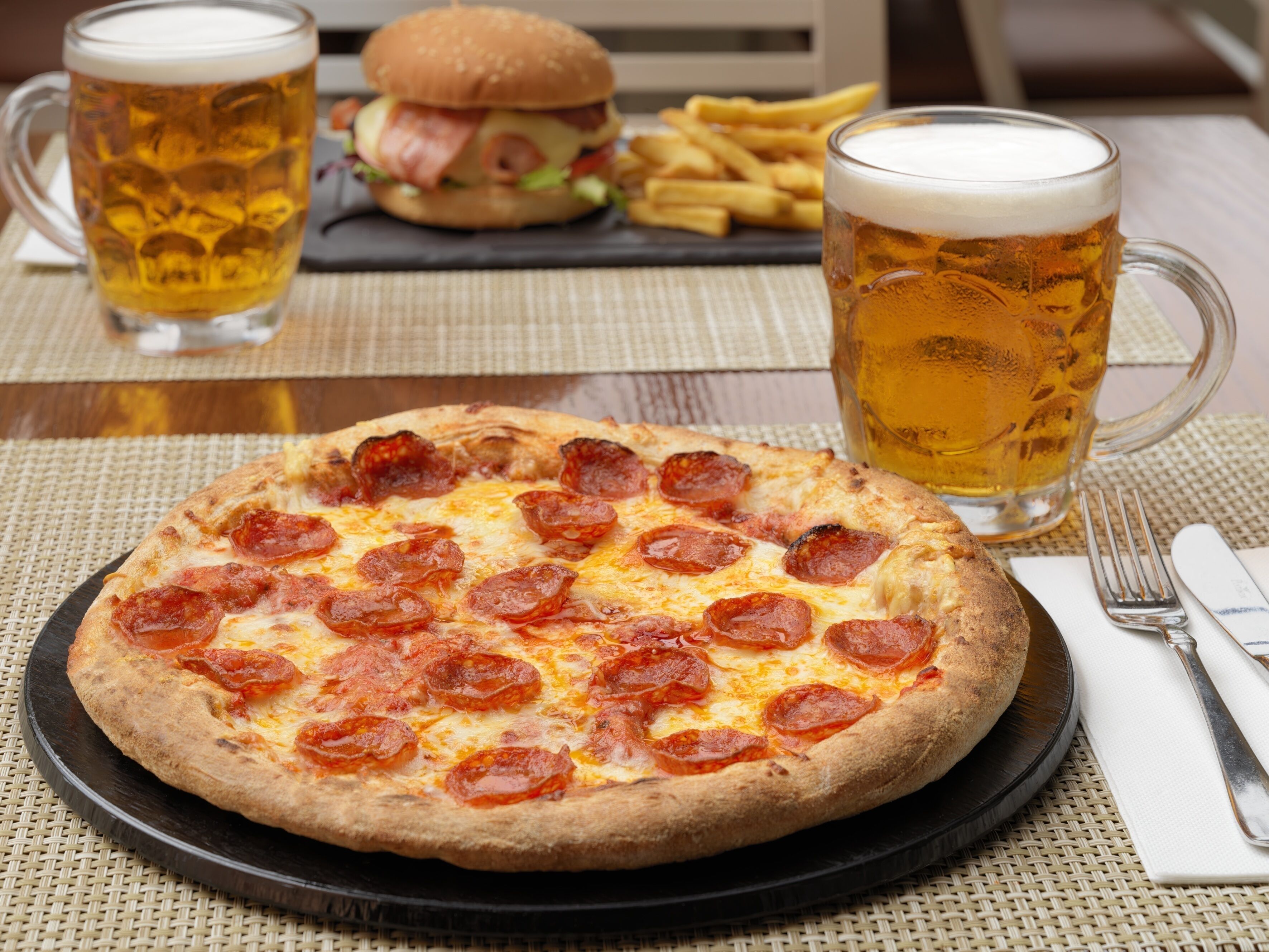 A pizza with pepperoni, a burger, fries, and two beers on a table.