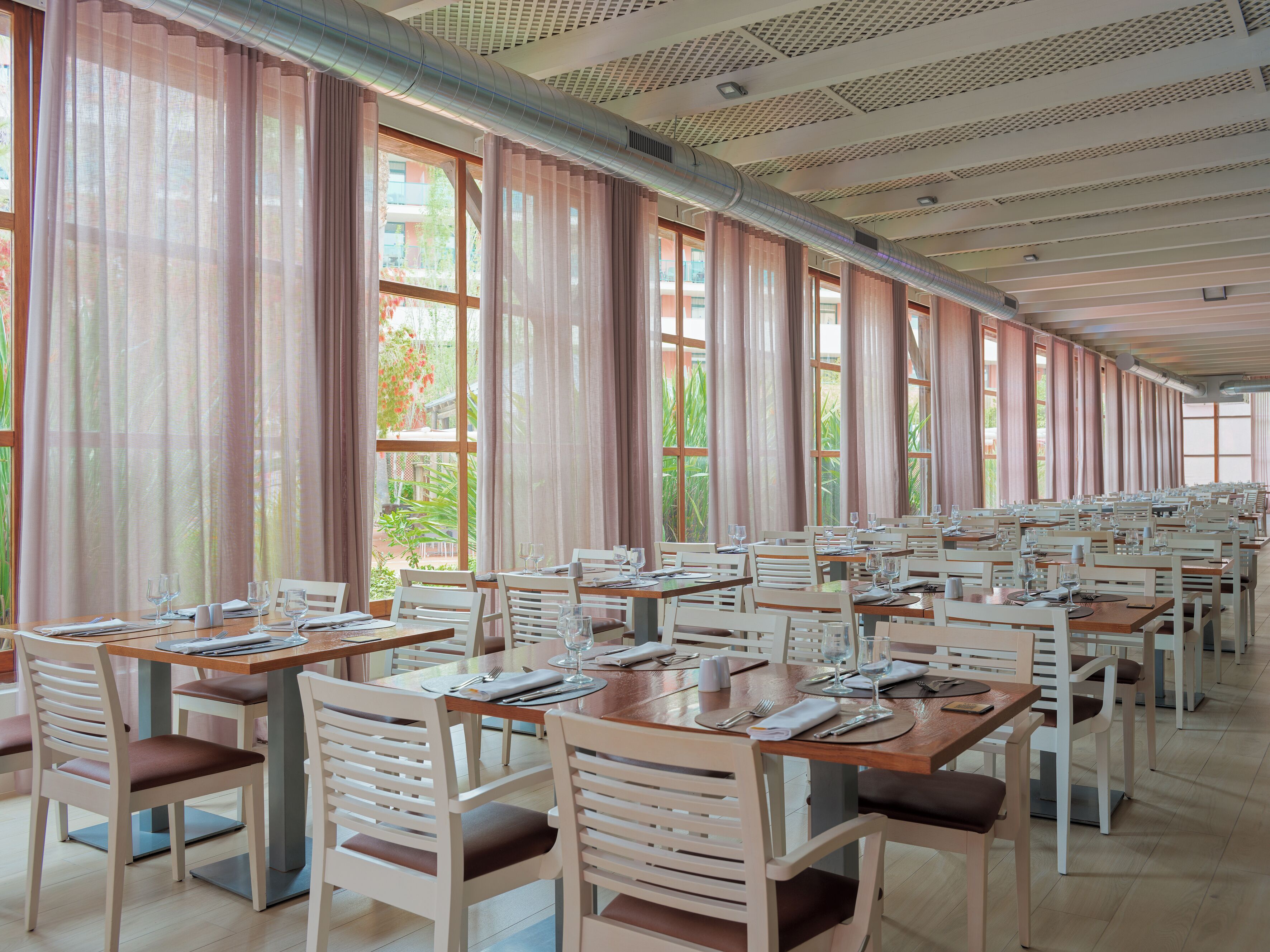Un comedor amplio con mesas y sillas largas, ventanas grandes con cortinas y un techo alto con conductos de ventilación.