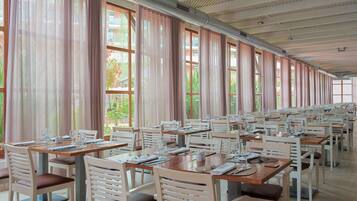 Un comedor amplio con mesas y sillas largas, ventanas grandes con cortinas y un techo alto con conductos de ventilación.