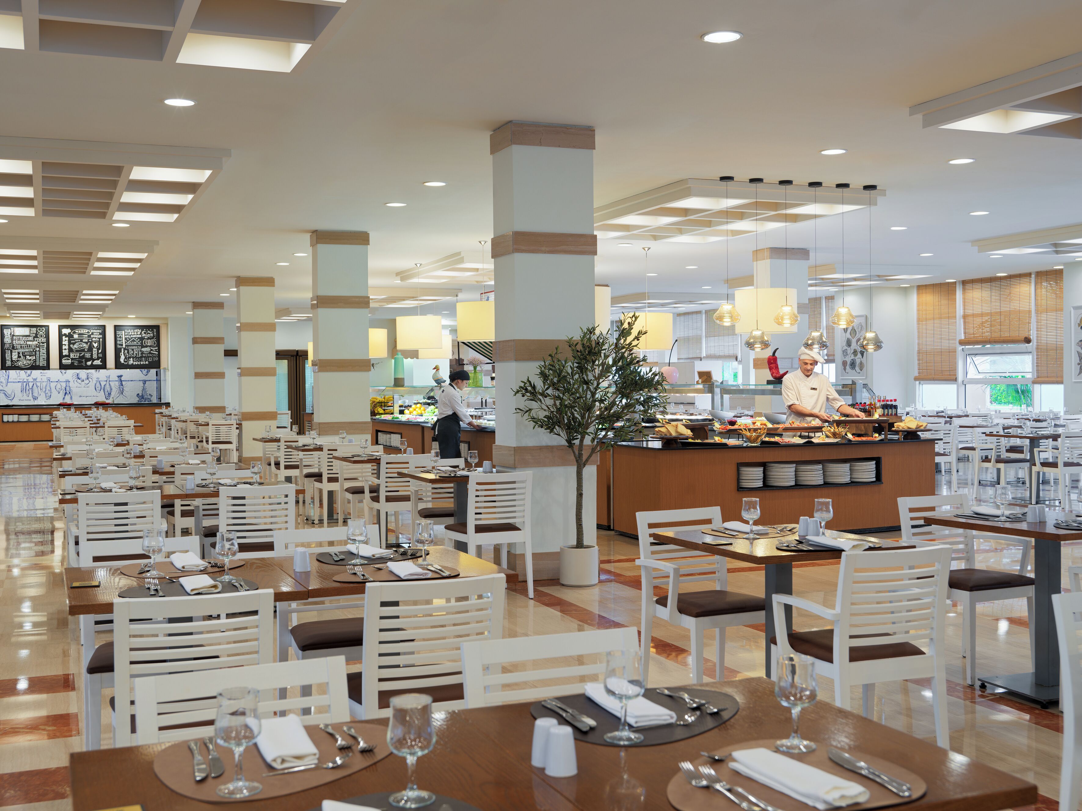 Un comedor amplio con múltiples mesas y sillas, un bufé central y un chef preparando comida.