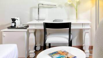 Habitación de hotel con un escritorio blanco, una silla, una mesita de noche con cafetera, una lámpara y una revista sobre una mesa redonda.