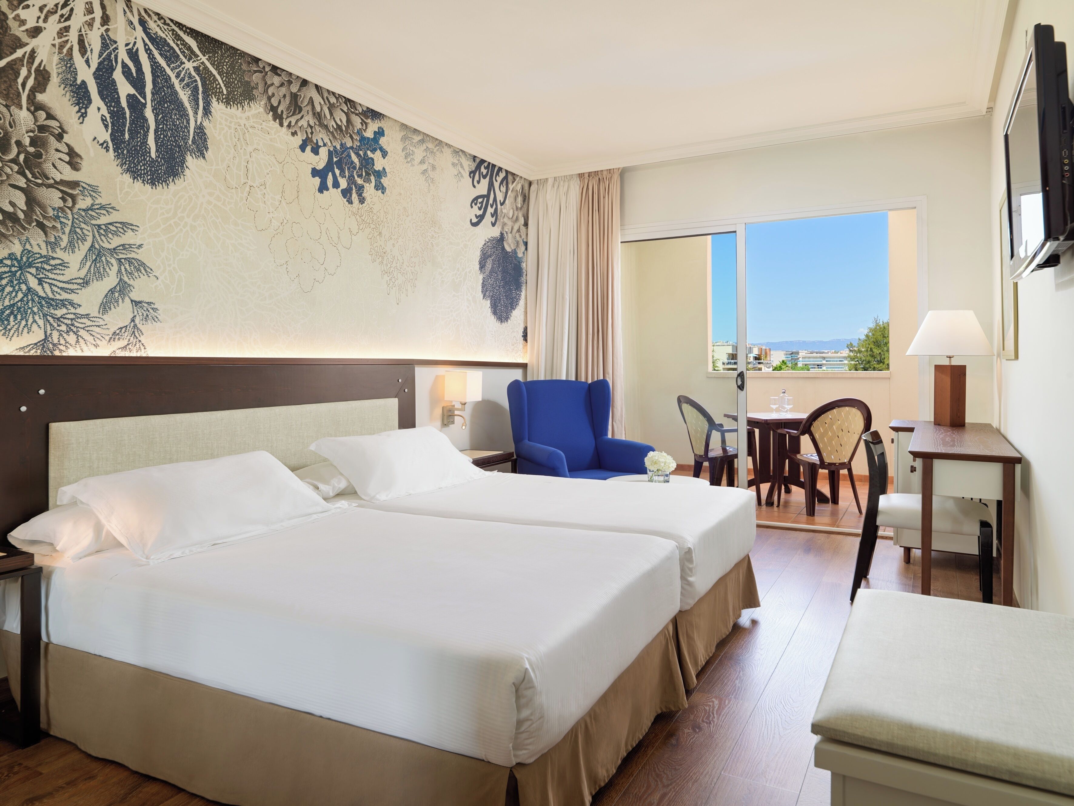 A hotel room with two beds, a blue armchair, a small table with a lamp, and a view of the sea through a large window.