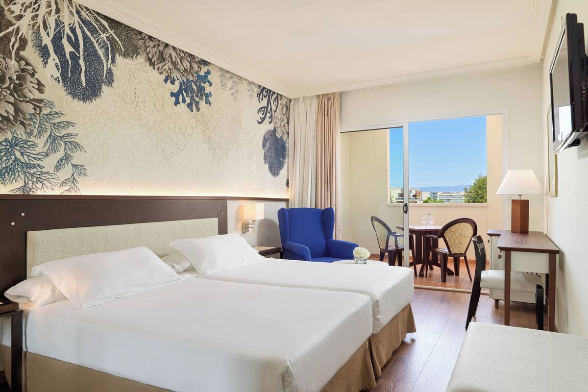 Une chambre d’hôtel avec deux lits, un fauteuil bleu, une petite table avec une lampe et une vue sur la mer par une grande fenêtre.