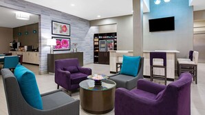 Een moderne hotellobby met paarse en grijze banken, een koffieapparaat, een minibar gevuld met drankjes en een flatscreen televisie aan de muur.