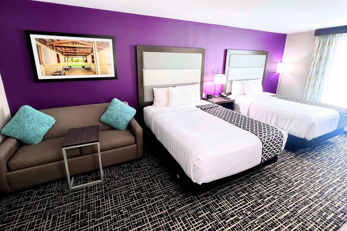 Une chambre d’hôtel avec deux lits, un canapé, un bureau et un mur de couleur violette.