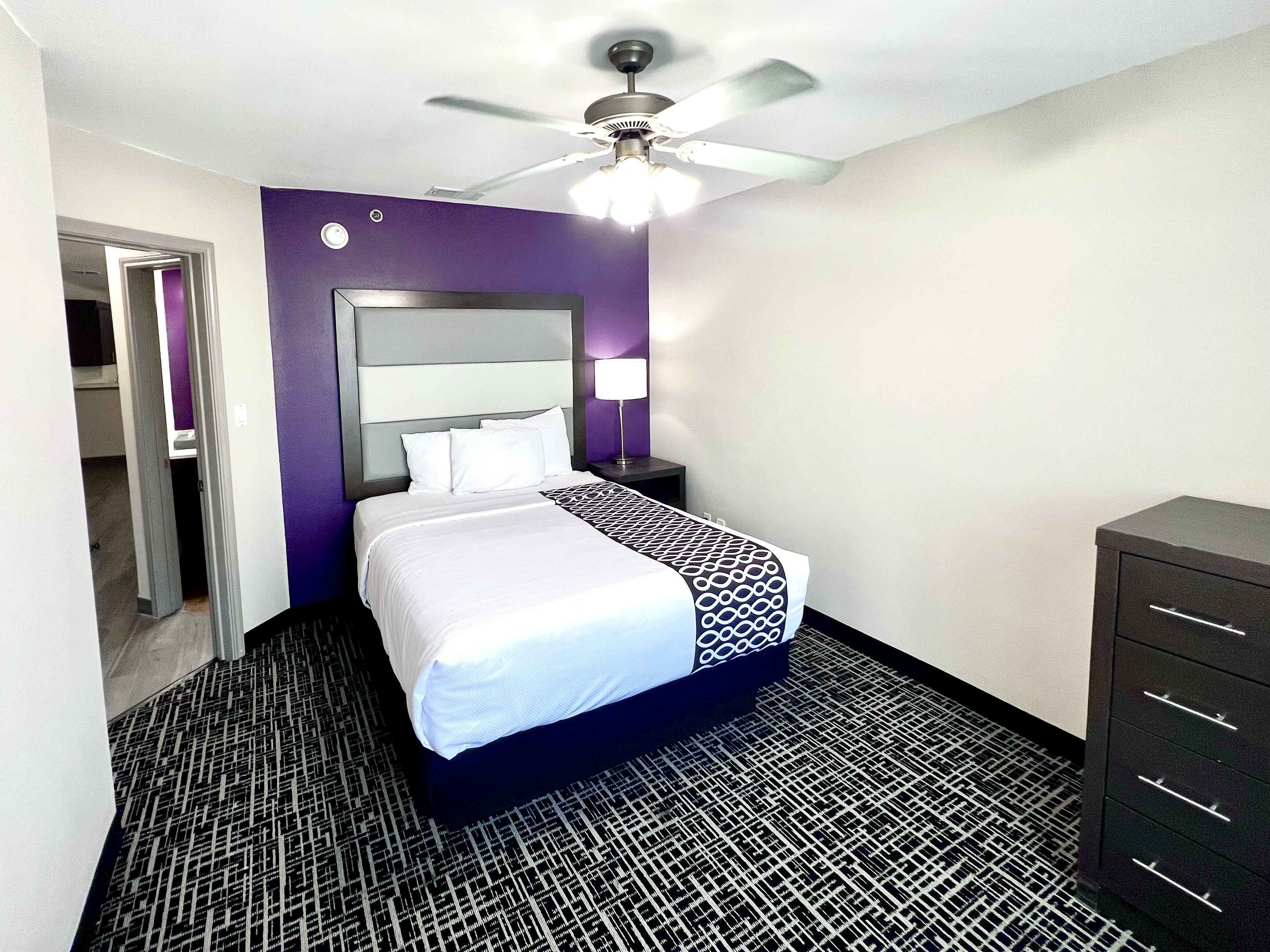Een hotelkamer met een bed, een plafondventilator, een nachtkastje en een dressoir.