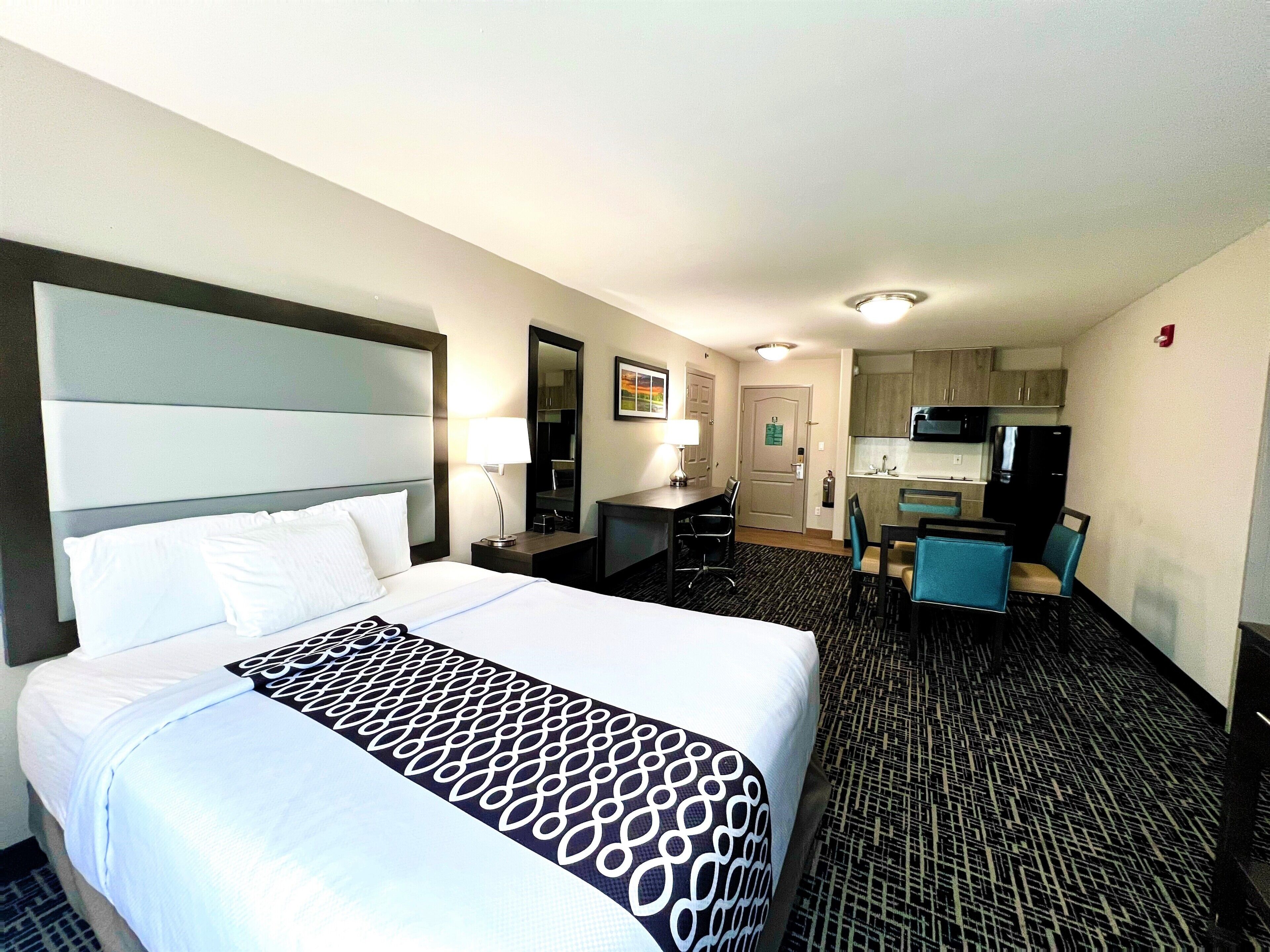 A hotel room with a large bed, a desk, a chair, a small kitchenette, and a bathroom door.
