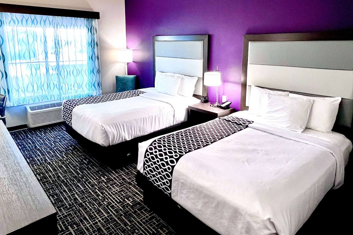 Une chambre d’hôtel avec deux lits, une fenêtre avec des rideaux, une table de chevet avec une lampe et un miroir fixé au mur.