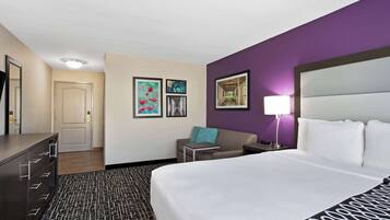 A hotel room with a bed, a desk, a chair, and a sofa. There are framed pictures on the wall.