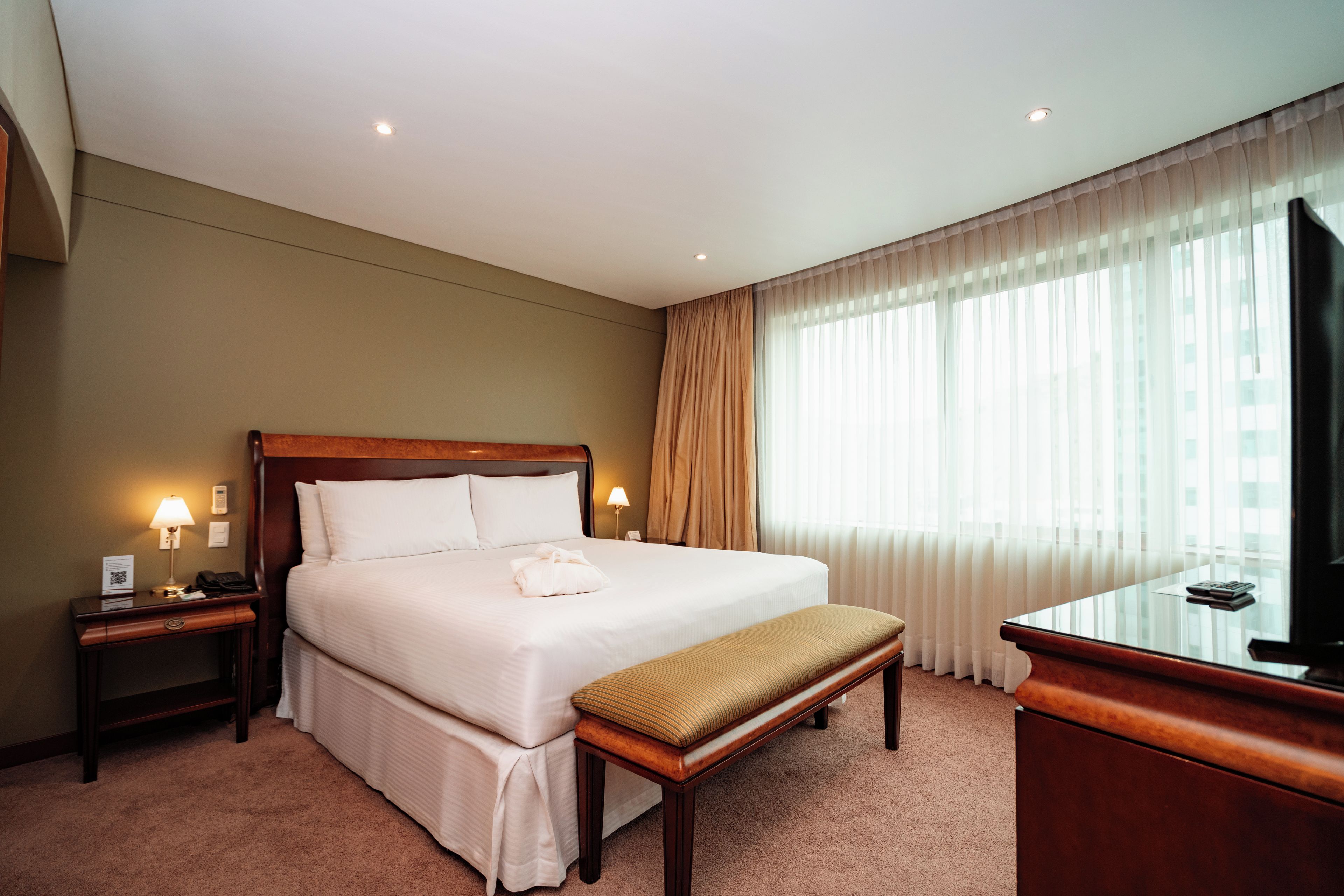 Una habitación de hotel con una cama grande, lámparas de noche, un escritorio con televisor y una ventana con cortinas translúcidas.