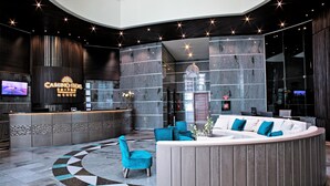 Un lobby moderno con un mostrador de recepción curvo, zona de estar con sillas color turquesa y un amplio muro de espejos.