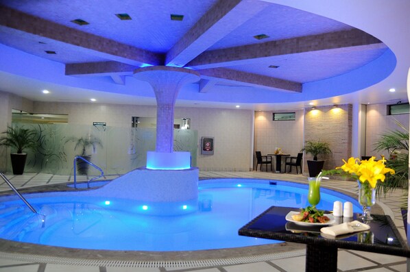 A swimming pool with a central fountain, surrounded by lounge chairs and tables, and a dining setup with a plate of food and drinks. - Suites Camino Real (La Paz)