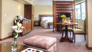 Una habitación de hotel con cama, una mesa de comedor con sillas, un sofá, una mesita con un jarrón y un marco de fotos, y un armario.