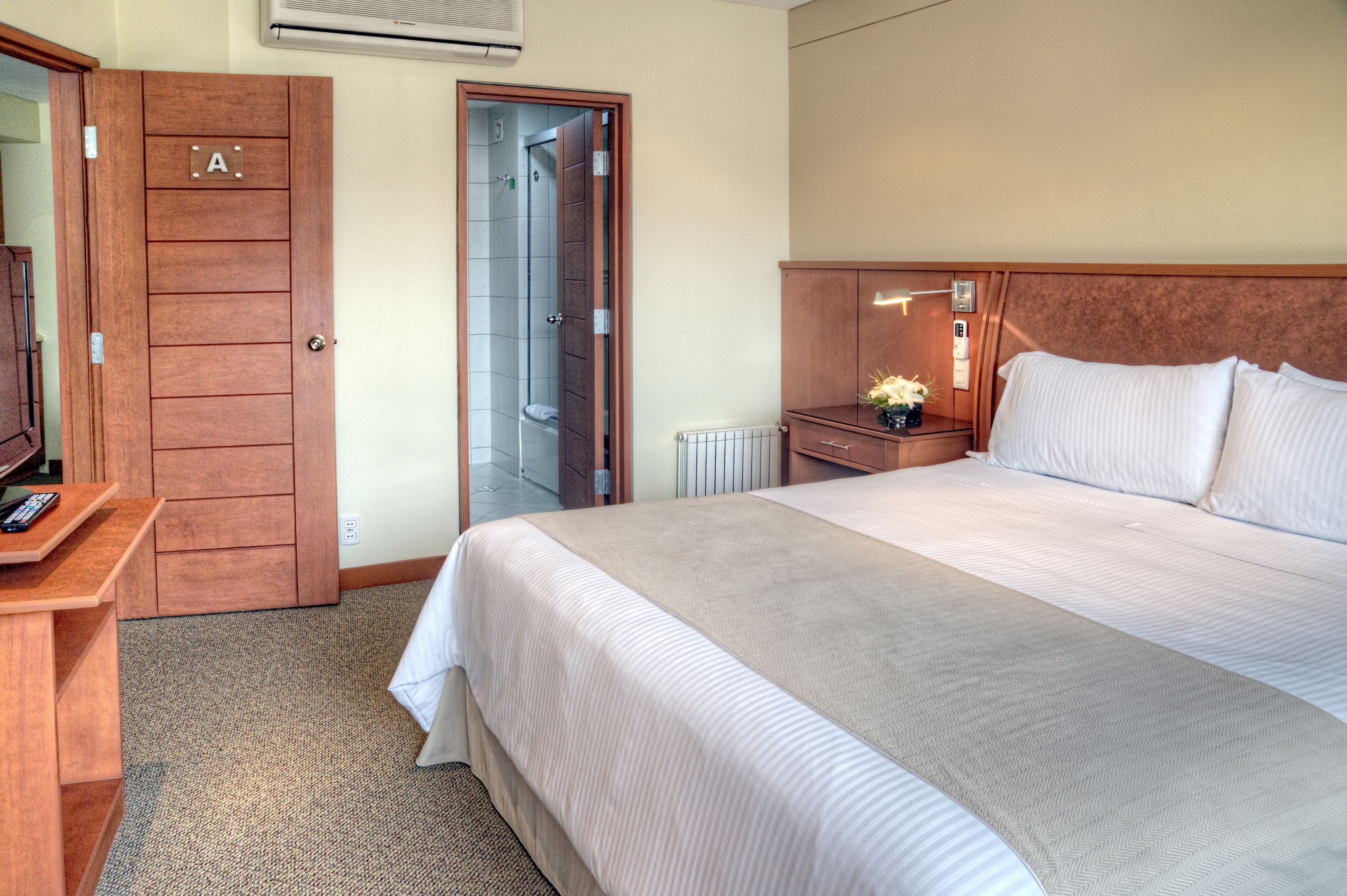 Habitación de hotel con una cama grande, un escritorio de madera con un control remoto, una mesita de noche con un florero, y un baño visible a través de una puerta abierta.