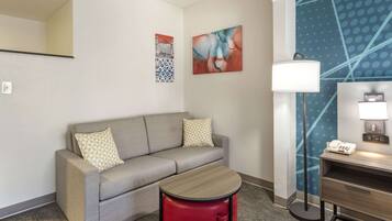 A hotel room with a grey sofa, a red trash bin, a wooden side table, a lamp, and a wall with abstract art.