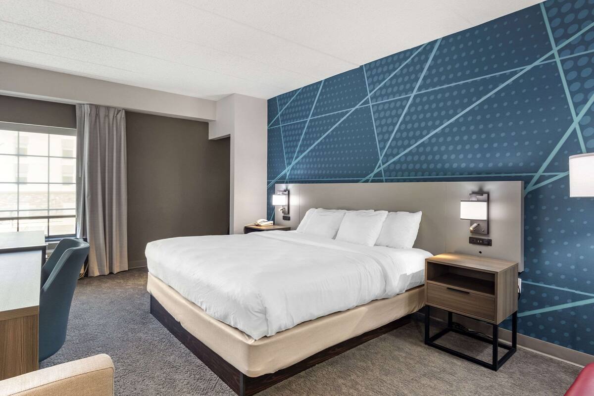 Habitación de hotel con una cama grande, un escritorio y una silla. Se ve una ventana con cortinas, una pared con un diseño geométrico y una mesita de noche con una lámpara.