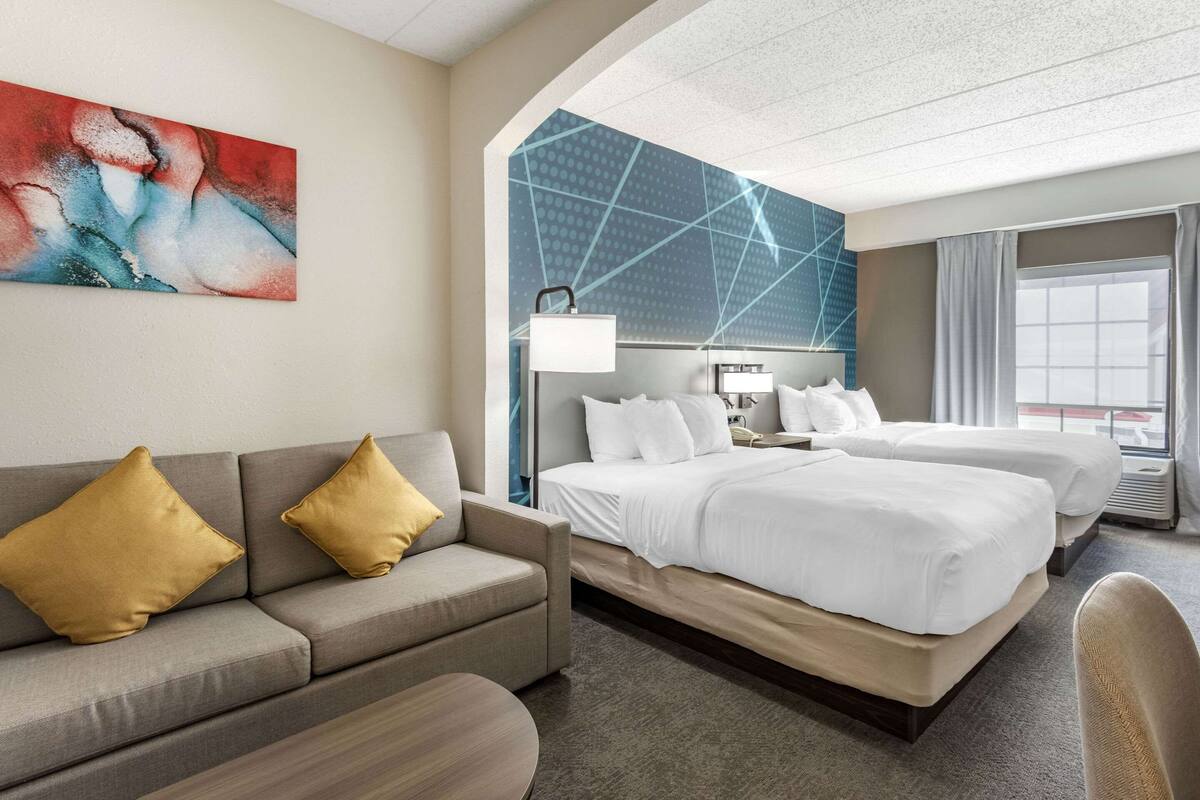 Una habitación de hotel con sofá, dos camas, un cuadro y un ventanal grande.