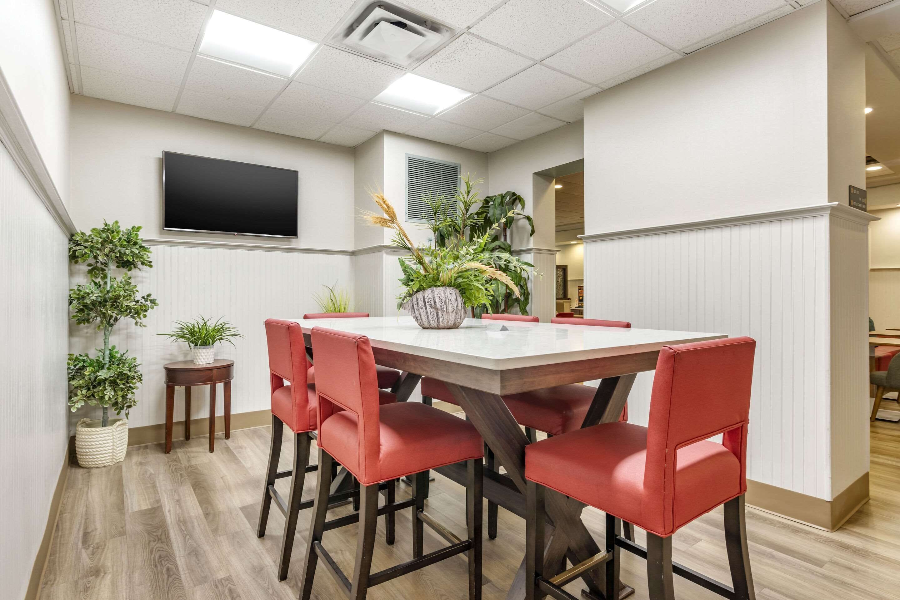 Un comedor con una mesa rectangular, seis sillas rojas y plantas en macetas.