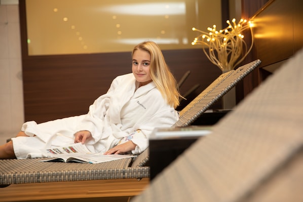 A person in a white bathrobe sitting on a wicker lounge chair in a room with a wooden floor and a decorative light fixture.