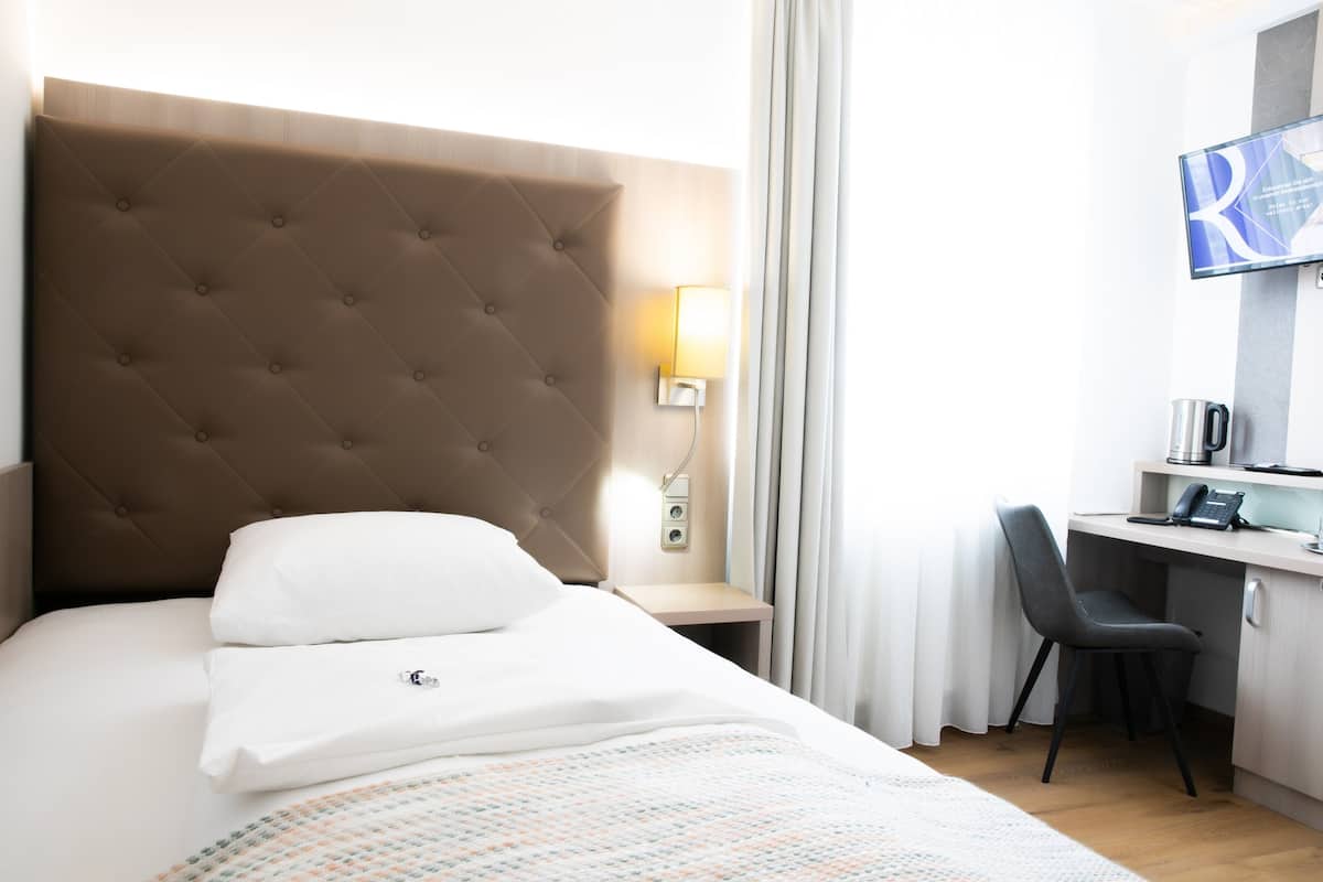 A hotel room with a tufted headboard, a desk with a chair, a flat-screen TV, and a bedside table with a lamp.