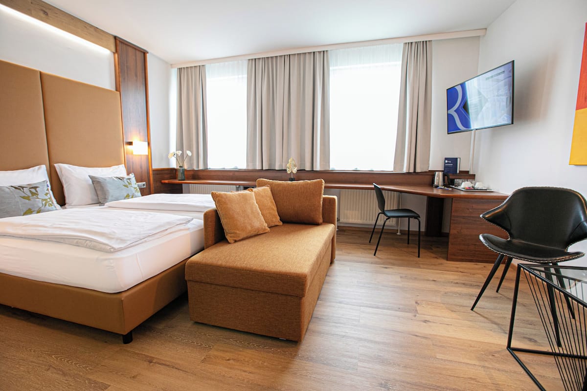 A hotel room with a bed, sofa, desk, chair, TV, and large windows.