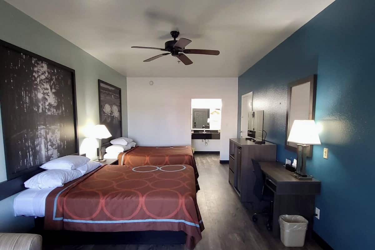 A hotel room with two beds, a ceiling fan, a desk with a lamp, a mirror, and a view of the bathroom.