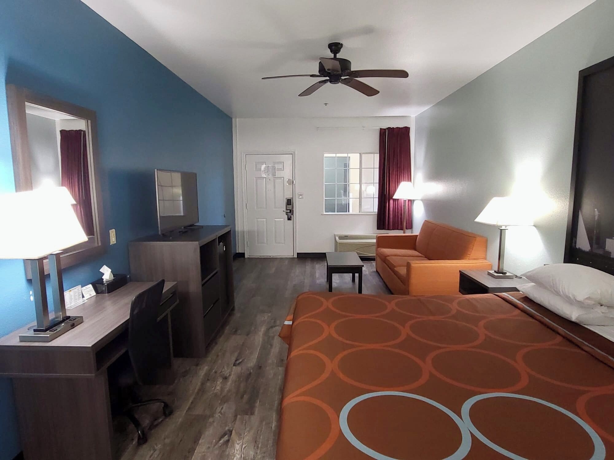 Habitación de hotel con cama, escritorio, silla, televisor, ventilador de techo y ventana con cortinas.